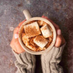 hands holding a cup of homemade coffee with marshmallows and cinnamon